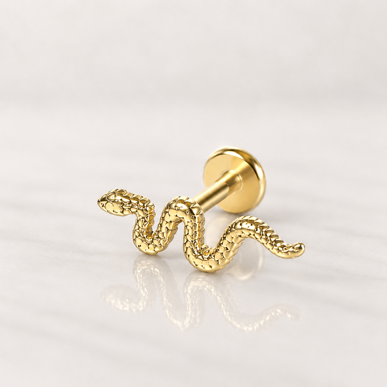 Labret doré à motif serpent aux écailles ciselées, base plate ronde, posé sur fond marbré clair