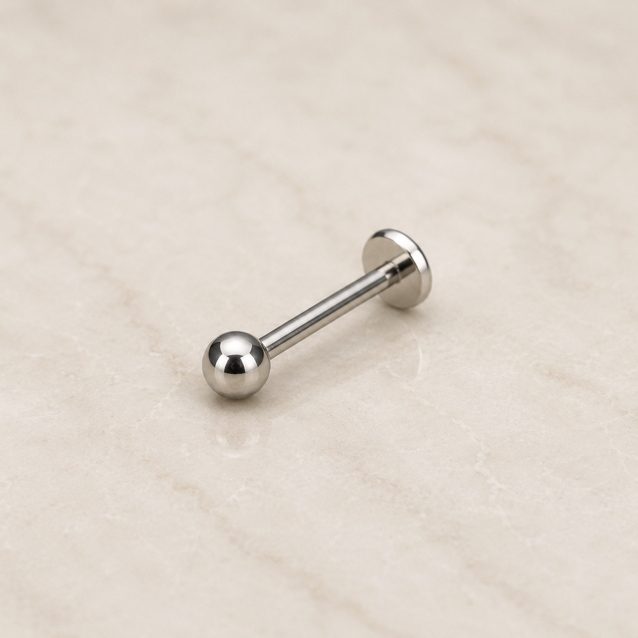 Labret en titane argente avec disque plat et boule ronde de 3 mm, pose sur un fond beige clair