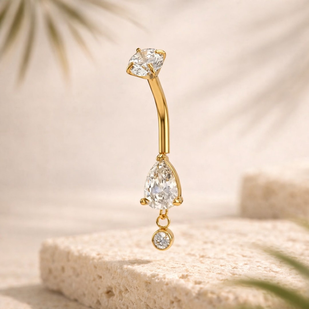 Banane de nombril dorée avec zircon rond, grande pierre en goutte et petit pendant serti, posée sur pierre claire