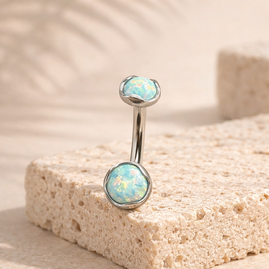 Banane de nombril en titane argenté avec deux cabochons bleu opalescent sertis à bord ondulé, posée sur une pierre claire