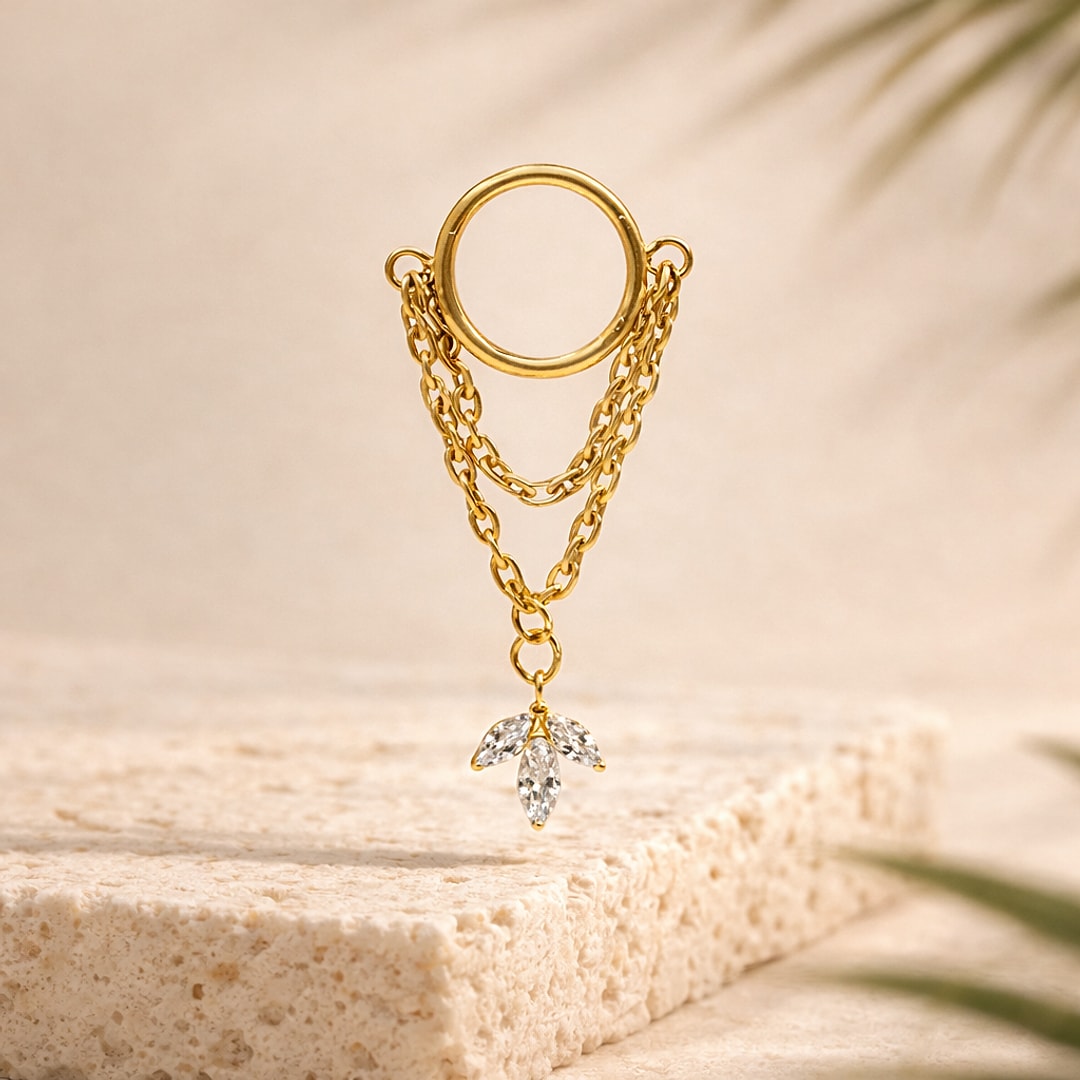 Anneau clicker en titane doré avec deux chaînes et pendentif feuille à trois zircons, posé sur pierre claire