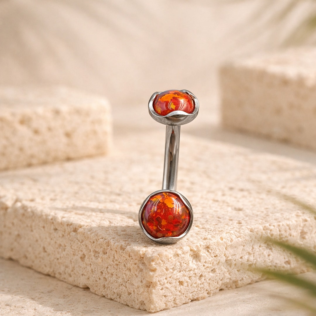 Banane de nombril en titane argenté avec deux cabochons rouge orangé sertis à bord ondulé, posée sur une pierre claire