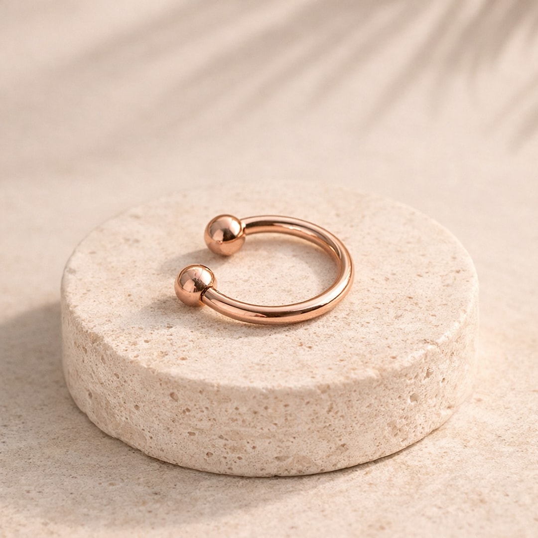 Fer à cheval de piercing en titane doré or rose avec deux boules, posé sur un socle de pierre claire