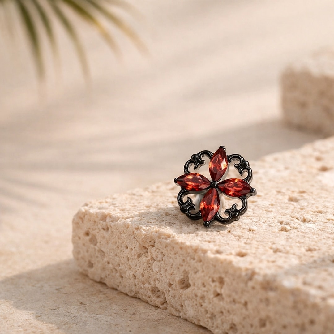 Stud flat back en titane noir orné d'une fleur baroque ajourée à quatre zircons rouges navette, posé sur une pierre claire