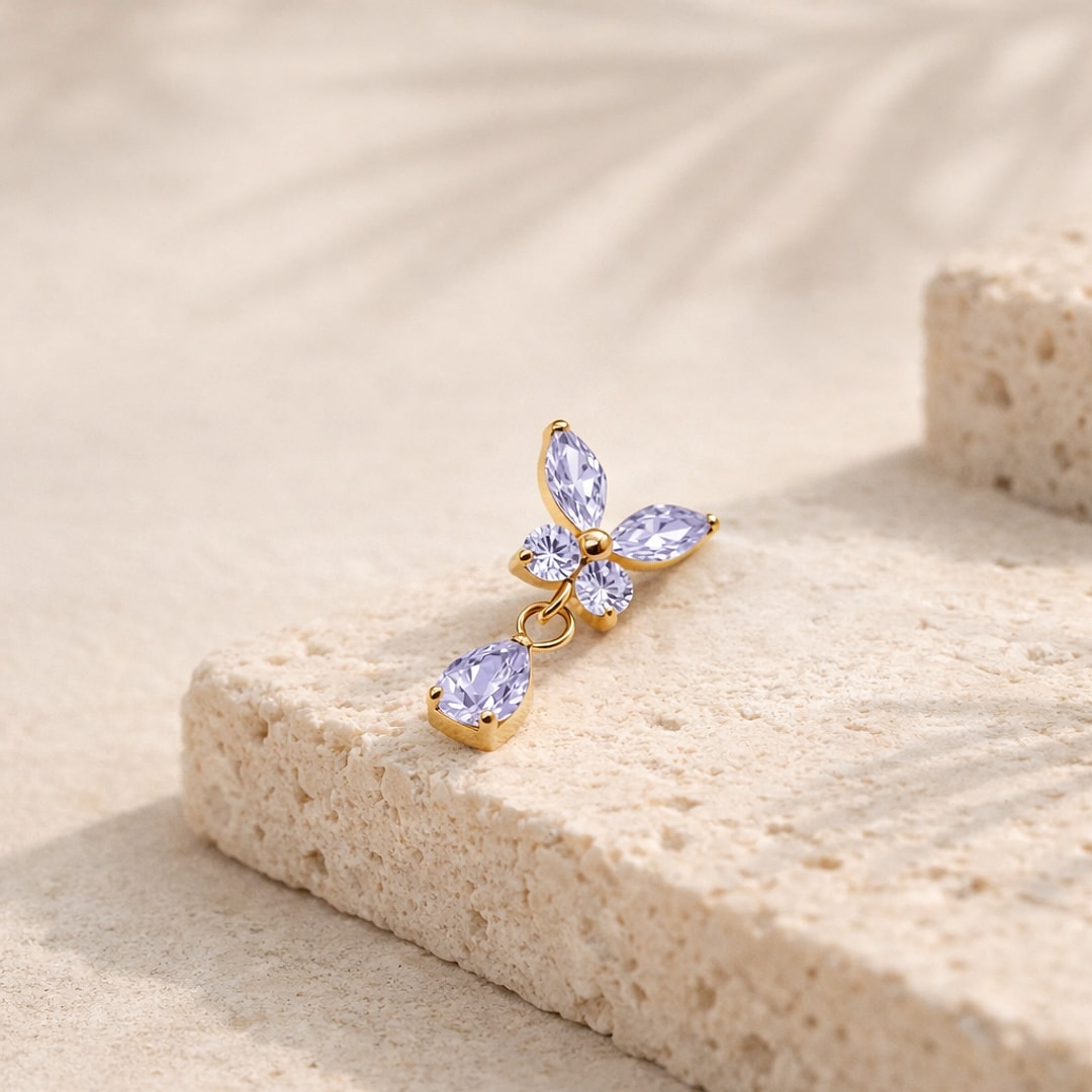 Top de piercing papillon en titane doré serti de zircons lilas avec goutte pendante, posé sur une pierre claire