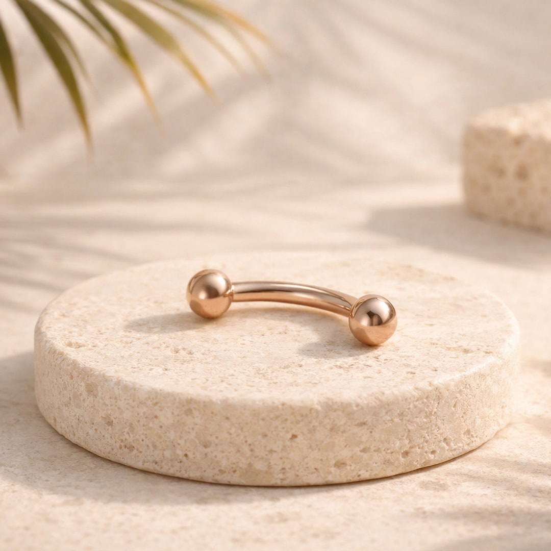 Barbell courbé en titane doré or rose à deux billes, posé sur un socle en pierre claire