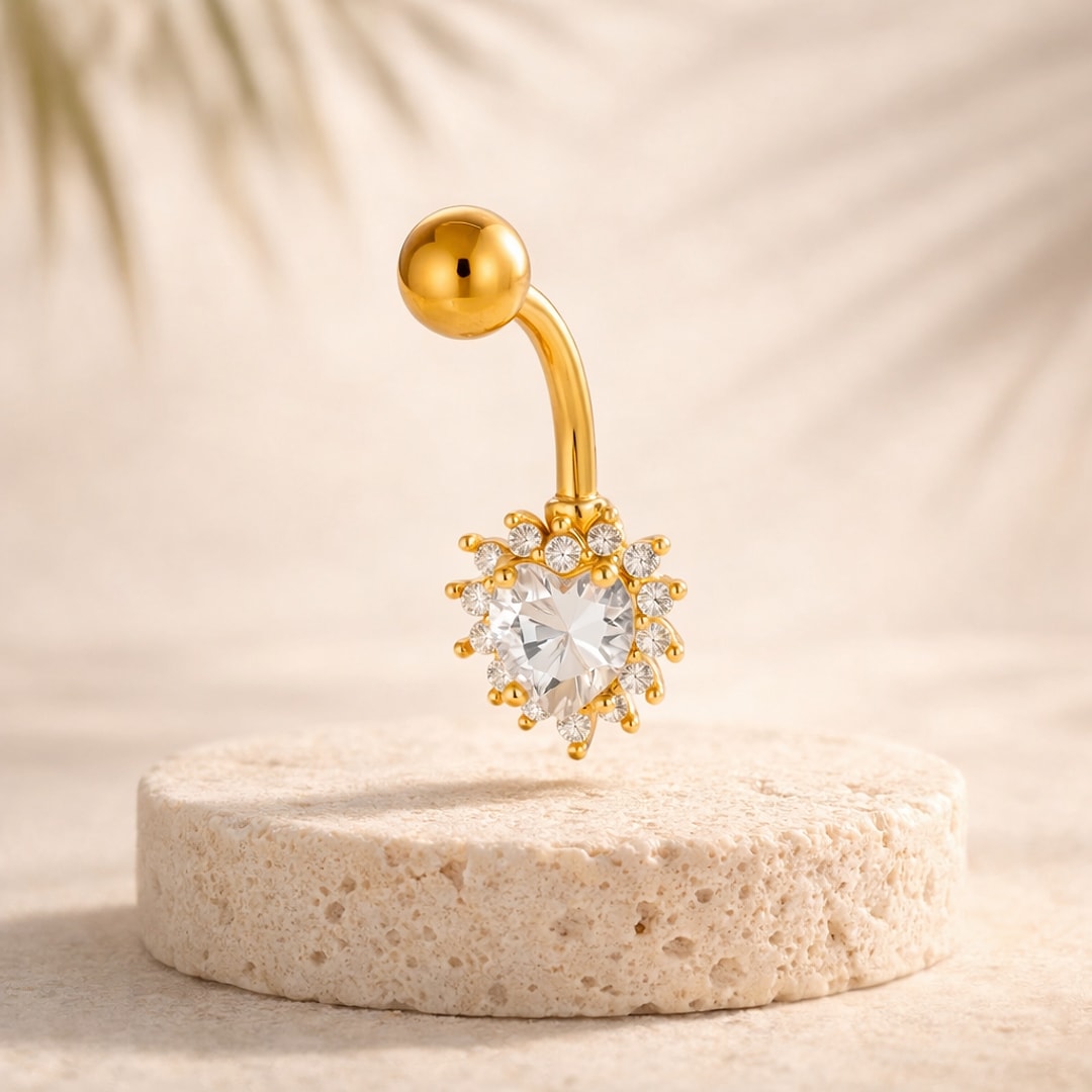 Banane de nombril doree avec coeur en cristal entoure de petites pierres, posee sur une pierre claire