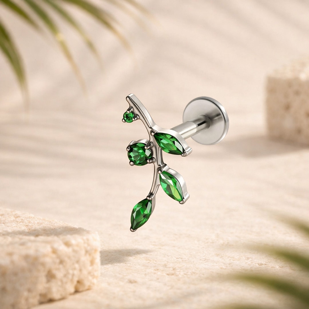 Stud flat back titane en forme de branche aux feuilles serties de zircons verts, posé sur du sable clair