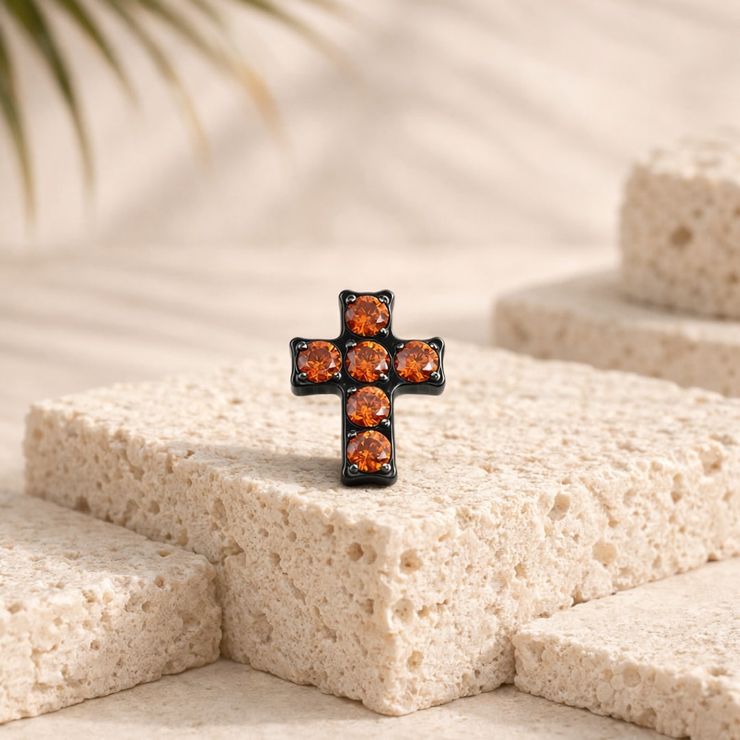 Embout de piercing croix en titane noir serti de six zircons orange, posé sur une pierre claire