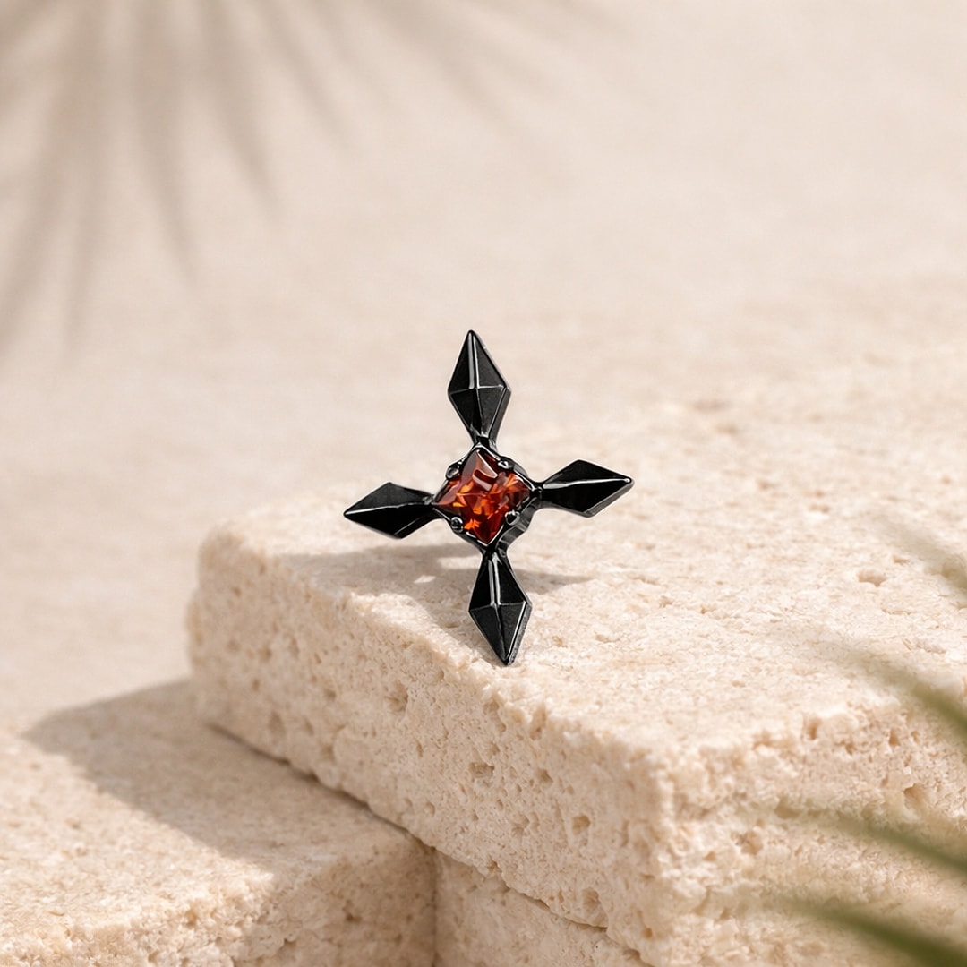 Stud flat back en titane noir en forme de croix à branches facettées, zircon rouge carré serti au centre, posé sur une pierre claire