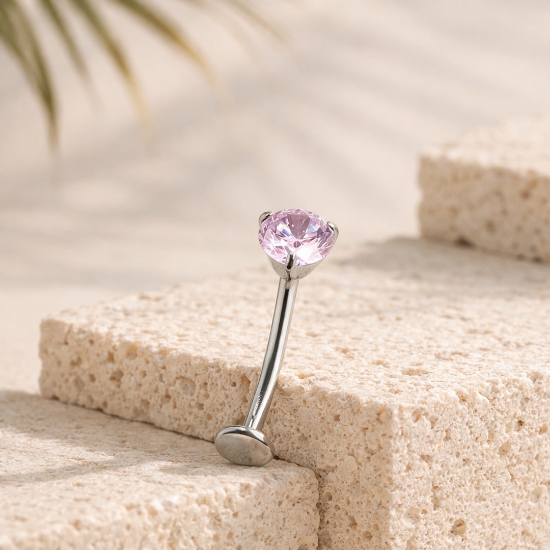 Banane de piercing nombril en titane argenté avec zircon rond rose serti griffes, posée sur une pierre claire