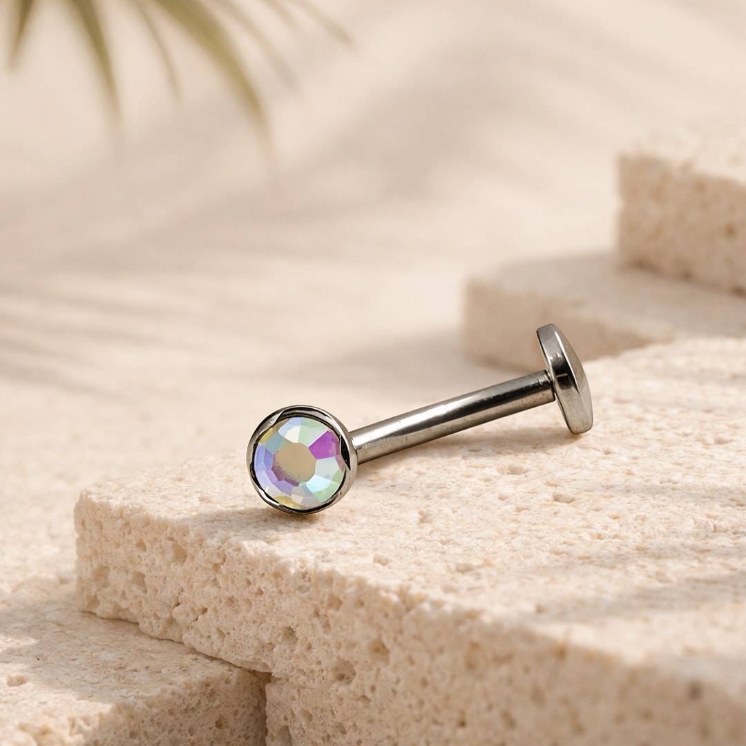Bijou de piercing en titane argenté à base plate, orné d'un cristal rond irisé vert et rose, posé sur une pierre claire