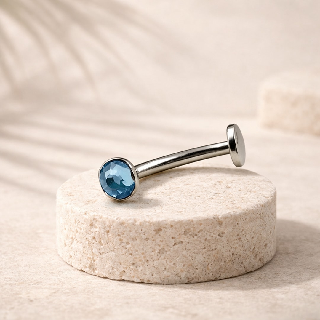 Banane de nombril en titane argenté avec cristal bleu facetté serti, posée sur un socle de pierre claire