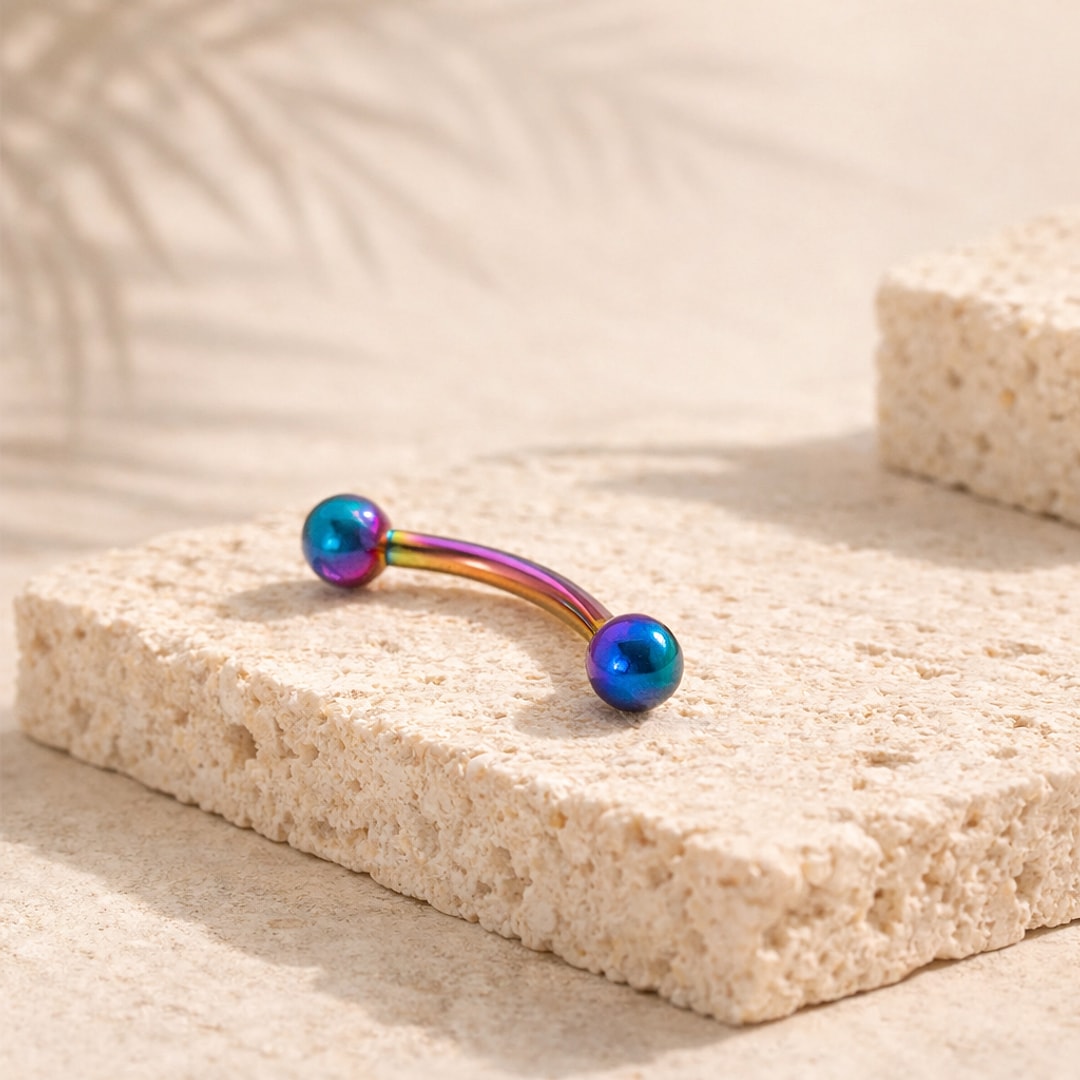 Barbell courbé en titane multicolore irisé avec deux billes, posé sur une pierre claire