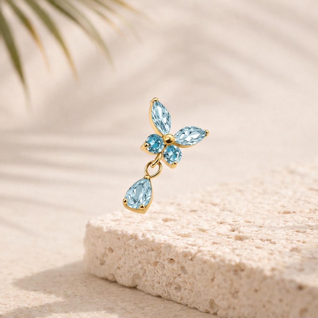 Top piercing papillon en titane doré serti de zircons bleus avec goutte pendante, posé sur une pierre claire