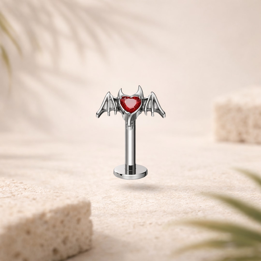 Stud flat back en titane argenté, cœur en zircon rouge entouré d'ailes de démon, sur fond beige