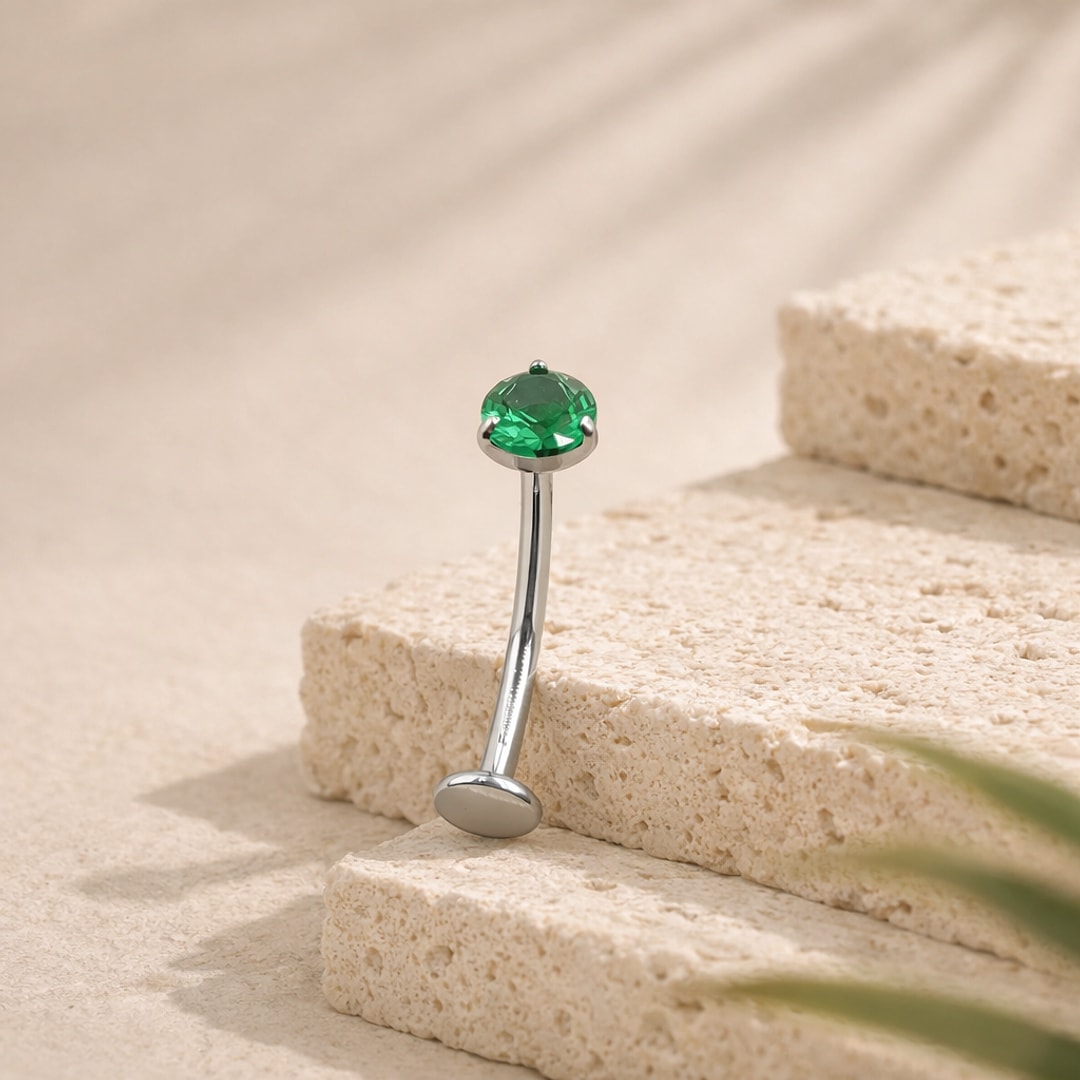 Banane de piercing nombril en titane argenté avec zircon rond vert serti griffes, posée sur une pierre claire
