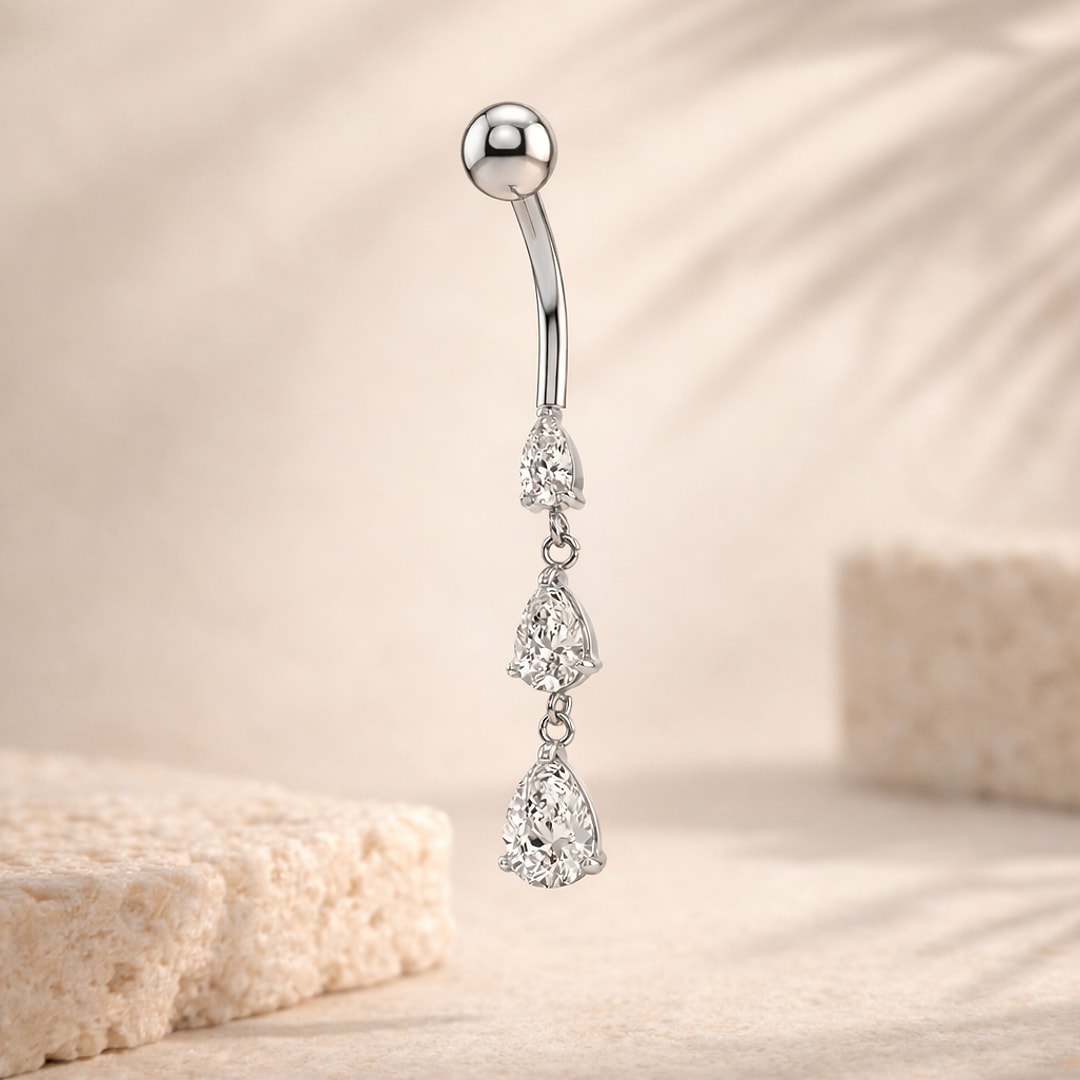 Piercing nombril acier argenté avec trois zircons blancs en forme de goutte pendants sur fond beige