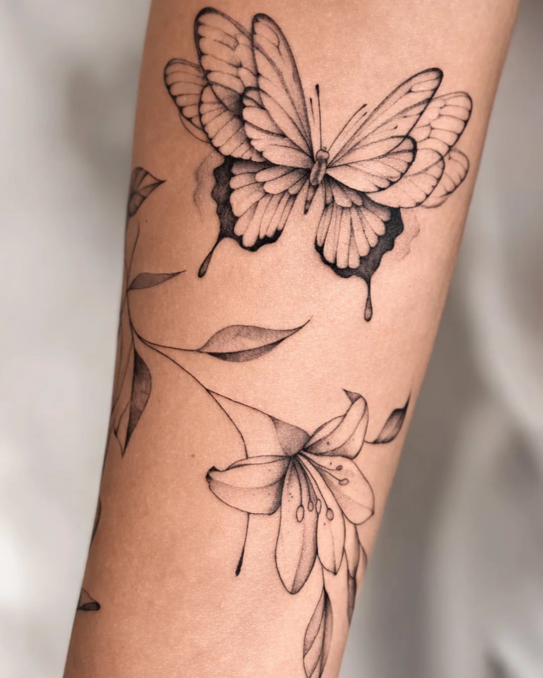 Tatouage papillon en fine line avec des fleurs de lys sur le bras, réalisé par Mathieu à Saint-Louis, La Réunion
