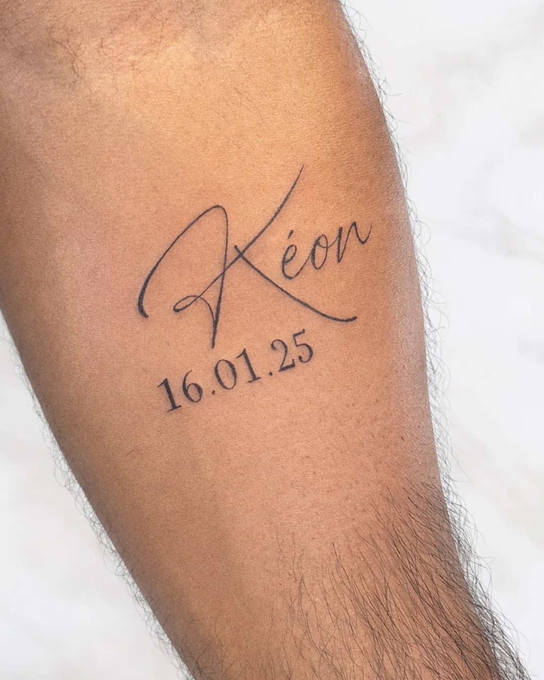 Tatouage prénom Kéon et date de naissance en écriture manuscrite sur l’avant-bras